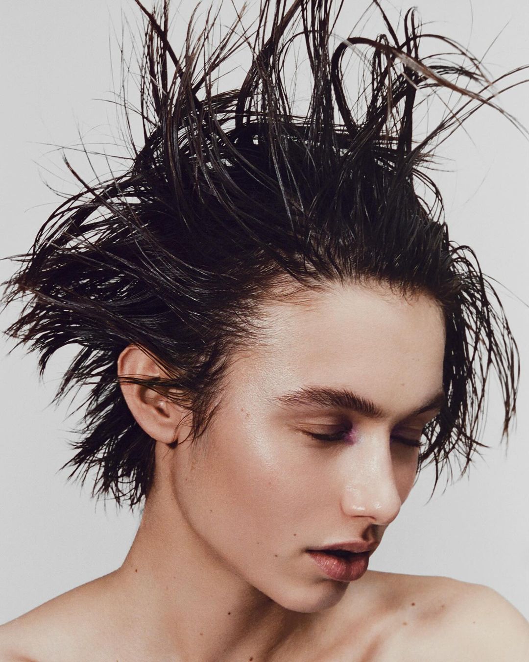 Close-up portrait of a young adult female with slicked-back dark hair, glossy skin, and closed eyes against a neutral background.