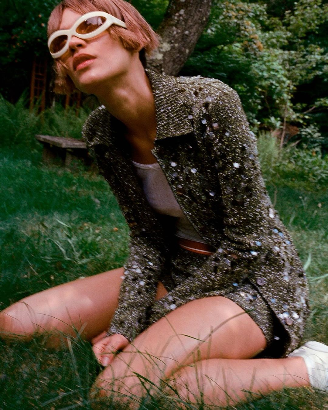 Young adult woman in a brown beaded jacket and shorts poses in a grassy outdoor setting with trees and dappled light, wearing sunglasses.