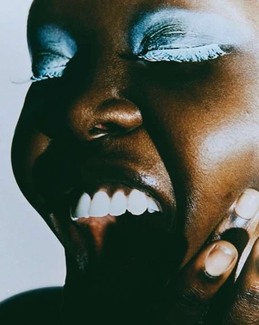Close-up portrait of a woman with blue eyeshadow and white eyelashes, showing an open mouth and manicured hand in a dimly lit setting.