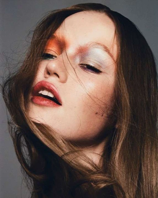 Beauty shot of a brunette woman with creative eye makeup, red lipstick, and wind-blown hair against a grey background.