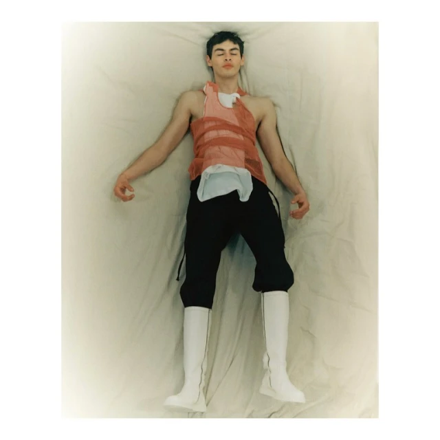 Overhead studio shot of a young adult man in high white boots lying on a white sheet wearing an orange mesh top and black pants.