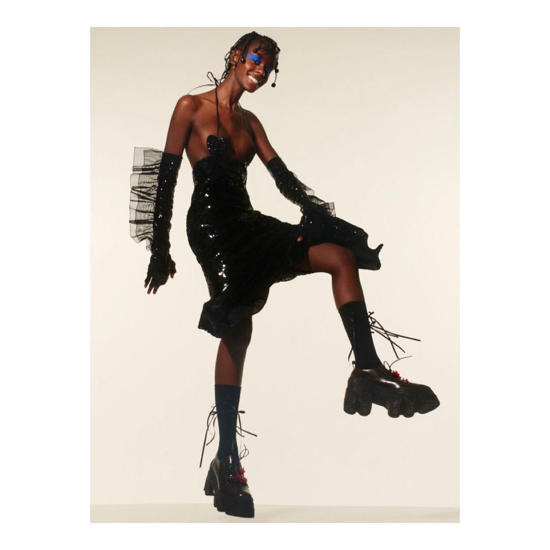A Black fashion model in a black sequin dress with gloves, posing indoors against a light backdrop, showcasing a fun and stylish look.