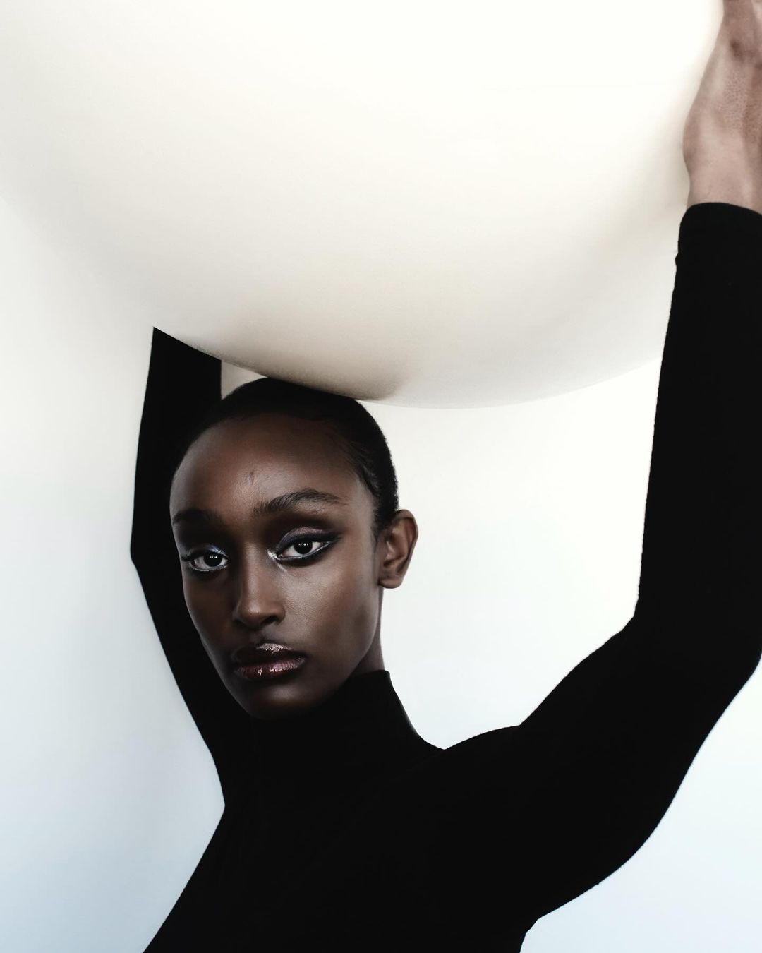 Portrait of a Black female model wearing a black turtleneck, striking makeup, and posing with arms raised against a stark white backdrop.