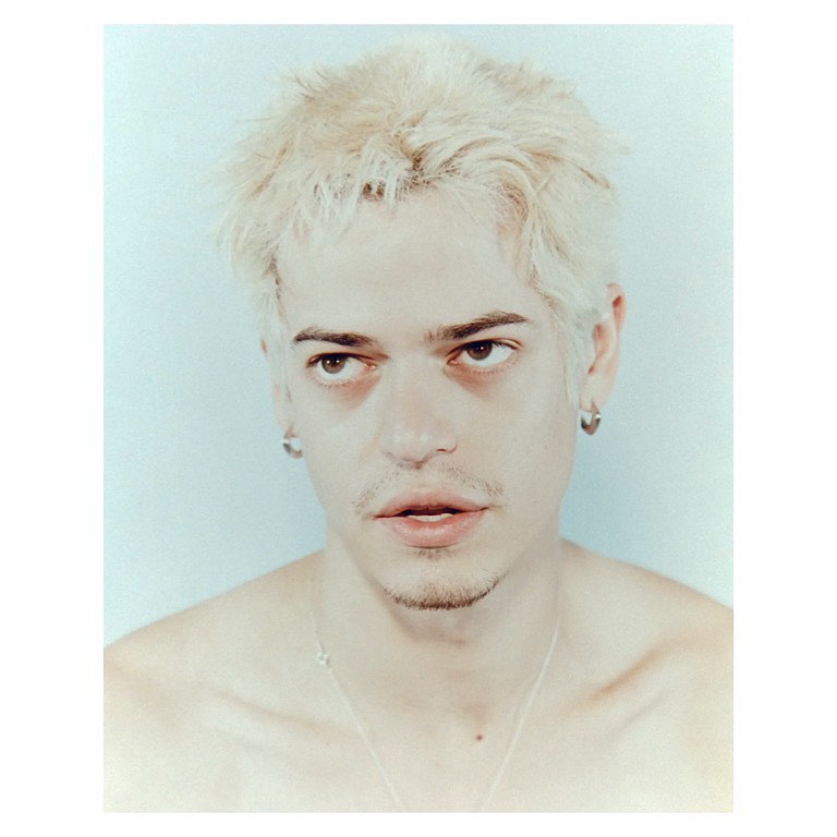 Studio portrait of a young man with blond hair and silver jewelry against a pale blue background with soft studio lighting.