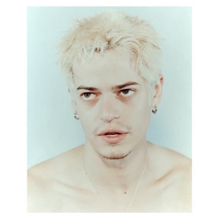 Studio portrait of a young man with blond hair and silver jewelry against a pale blue background with soft studio lighting.