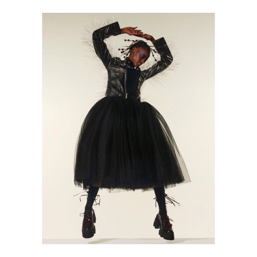 A striking editorial fashion shot of a Black woman in a black tulle dress, leather jacket, and platform shoes, creatively posed against a light background.