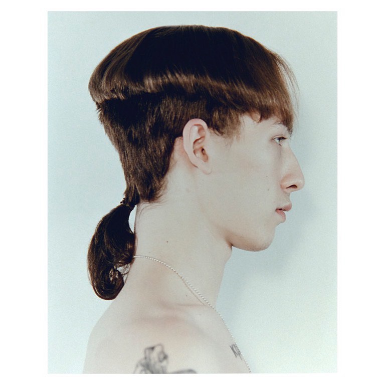 Profile shot of a bare-chested, pale-skinned young adult male with a unique dark brown hairstyle and visible tattoos against white backdrop.