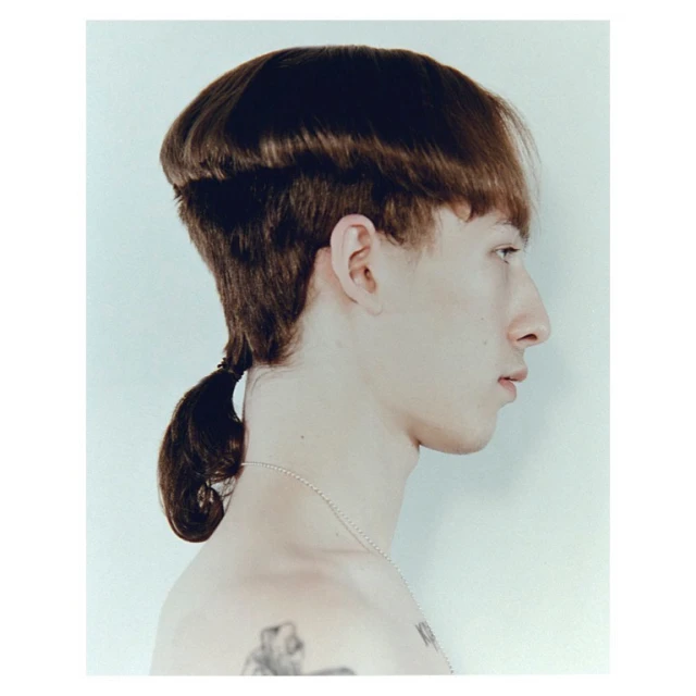 Profile shot of a bare-chested, pale-skinned young adult male with a unique dark brown hairstyle and visible tattoos against white backdrop.