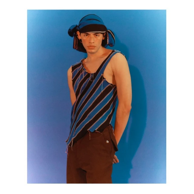 Stylish adult male poses in a fashion shot wearing a blue and black striped top and matching cap in front of a blue backdrop.