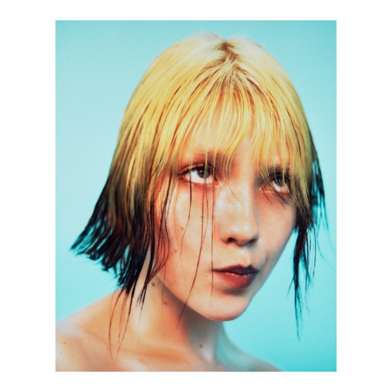 Close-up portrait of a young adult female with blonde and black hair, looking up against a light blue background in a studio setting.