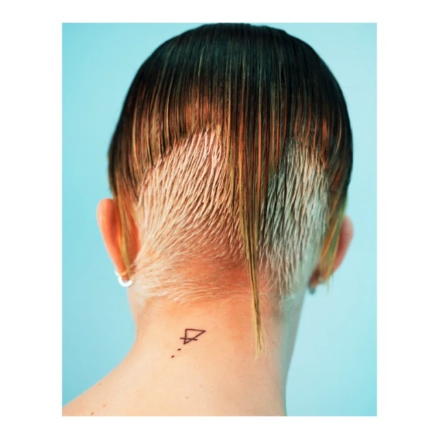 Back view of a person with a blonde undercut and a geometric tattoo on their neck against a light blue backdrop in a portrait shot.