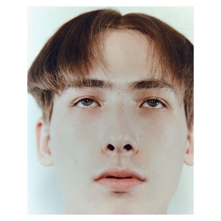 Close-up portrait of a young adult male with brown bowl cut hair and light skin tone against a plain light background.