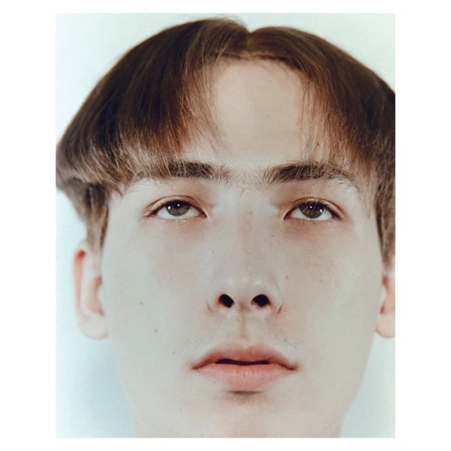 Close-up portrait of a young adult male with brown bowl cut hair and light skin tone against a plain light background.