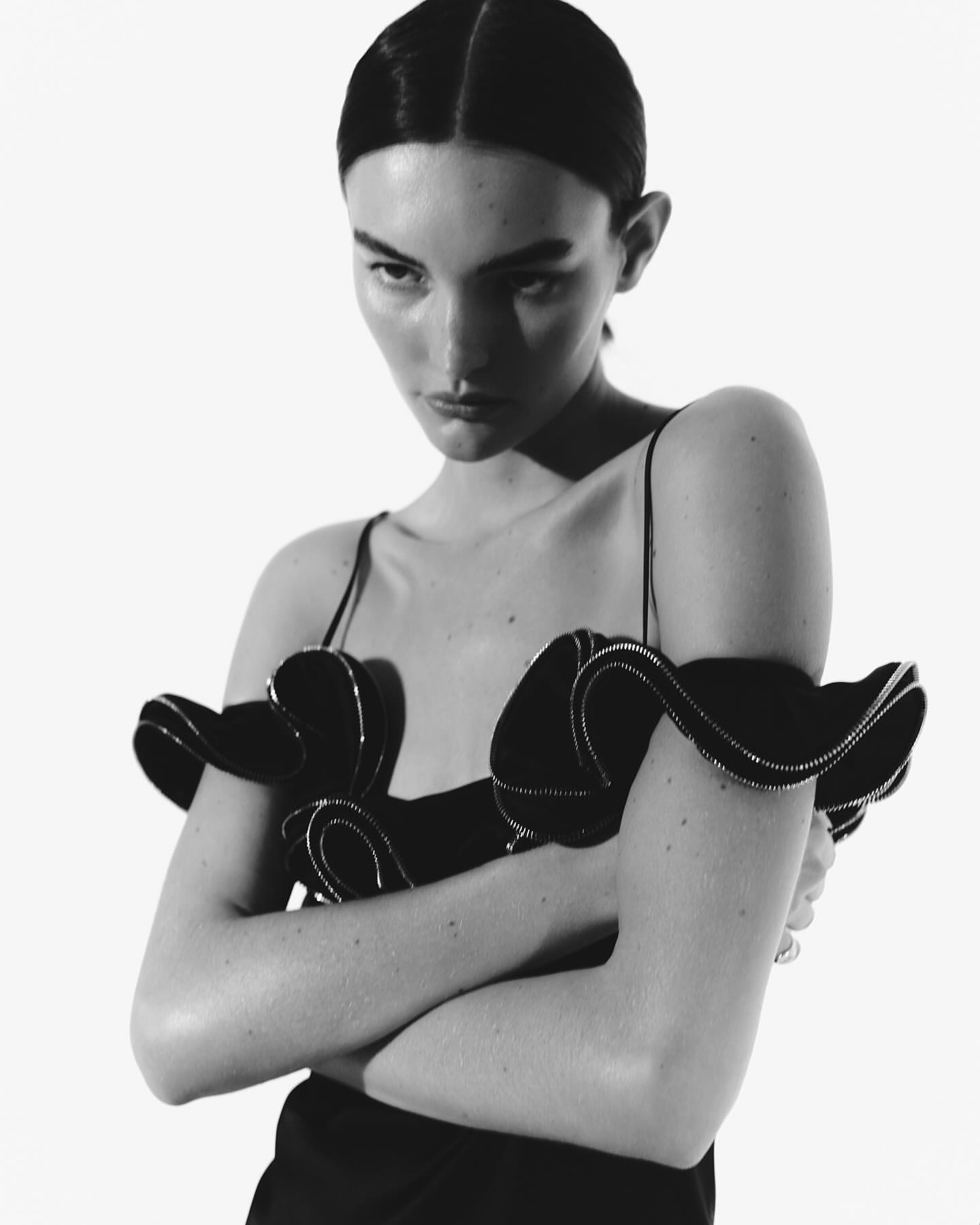 Black-and-white studio portrait of an adult woman with crossed arms wearing a ruffle-sleeved top against a bright white backdrop.