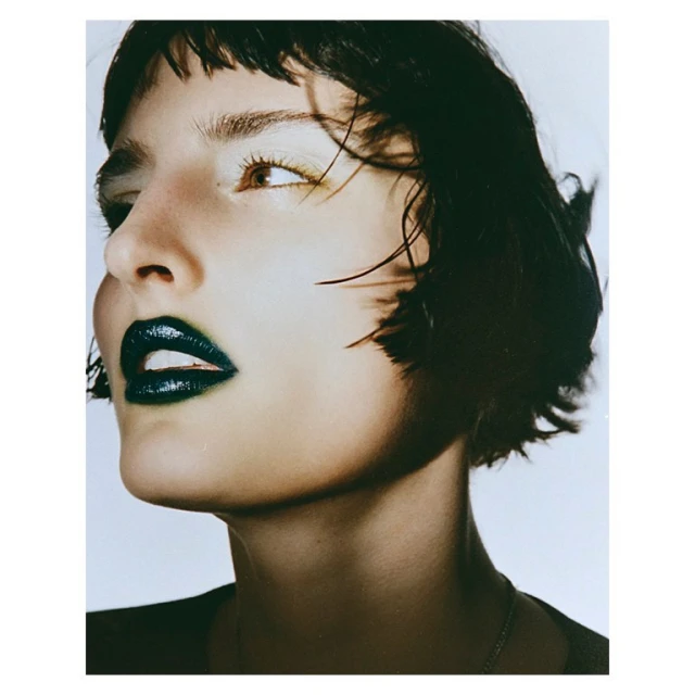 Close-up of a woman with short dark hair and dark, glossy lipstick looking up against a white background in a beauty style photograph.