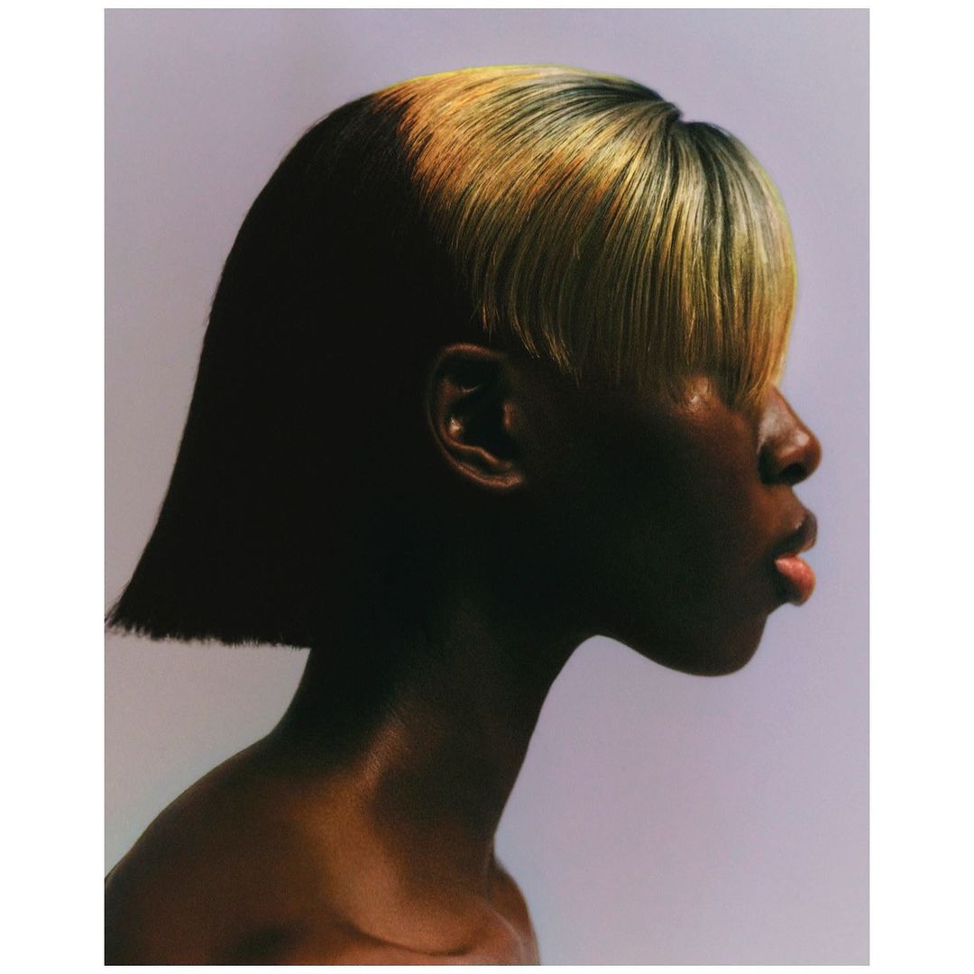 Close-up profile of an adult woman with dark skin and short blonde and black hair, against a light background in a studio shot.
