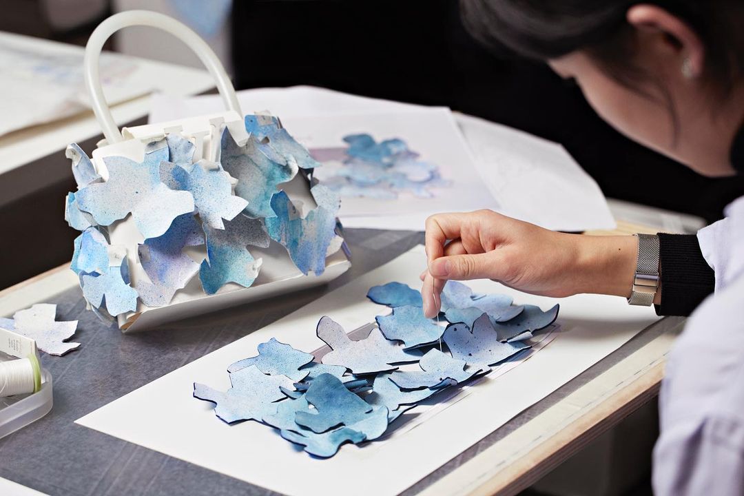Close-up of a white handbag covered with blue flower petals being assembled by a woman in a bright indoor setting, showcasing fashion craftsmanship.