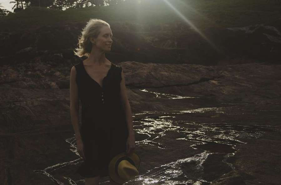 Blonde woman in a dark dress stands on rock formation near the coast at sunset holding a straw hat, with palm trees in background.