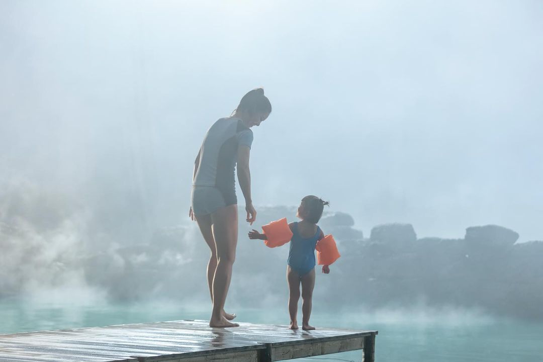 A mother and child stand at the edge of a misty lagoon, the little girl in arm floaties, creating a serene and heartwarming scene.