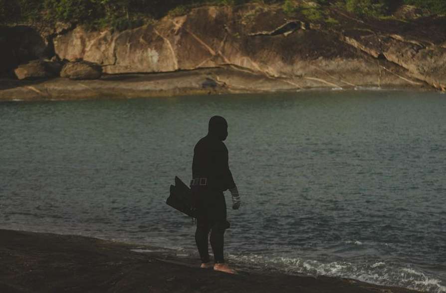 Silhouette of a diver in a wetsuit standing on a rocky shore with flippers and a mask, ocean and cliffs in the background.