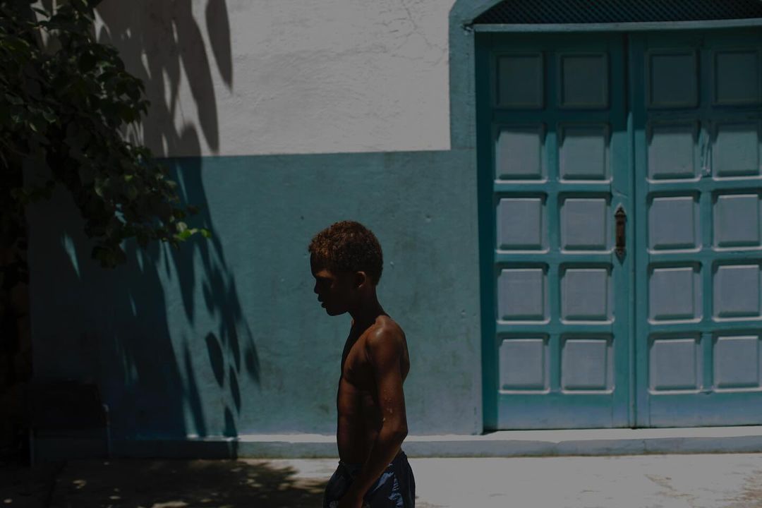 A young boy with dark skin and short, brown hair stands in profile view outside a building with teal doors. Sunlight and shadows.