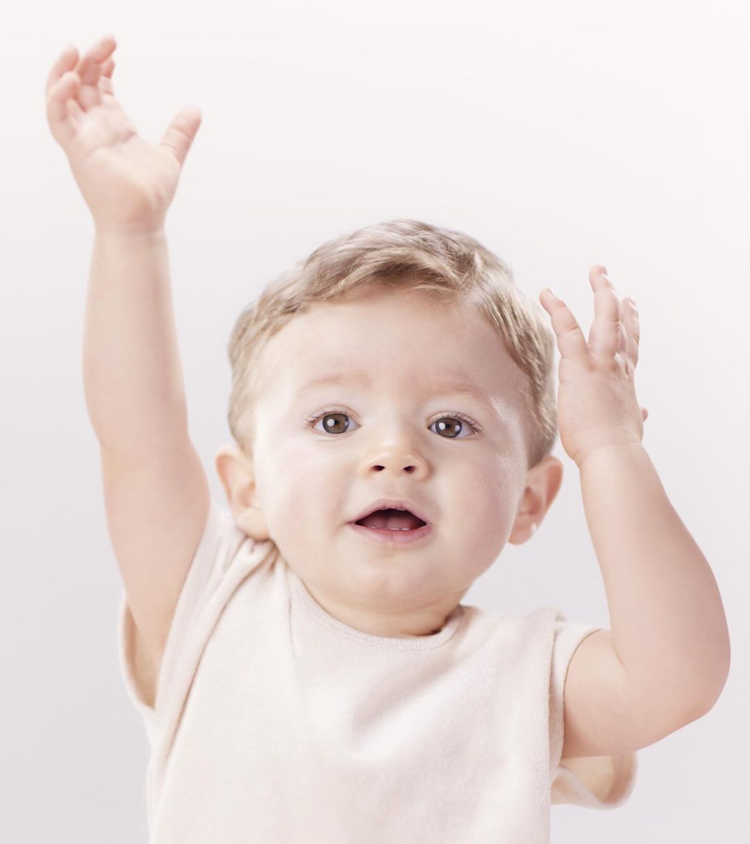 A baby with light brown hair smiles at the camera with arms raised against a neutral background. The baby wears a cream-colored shirt.
