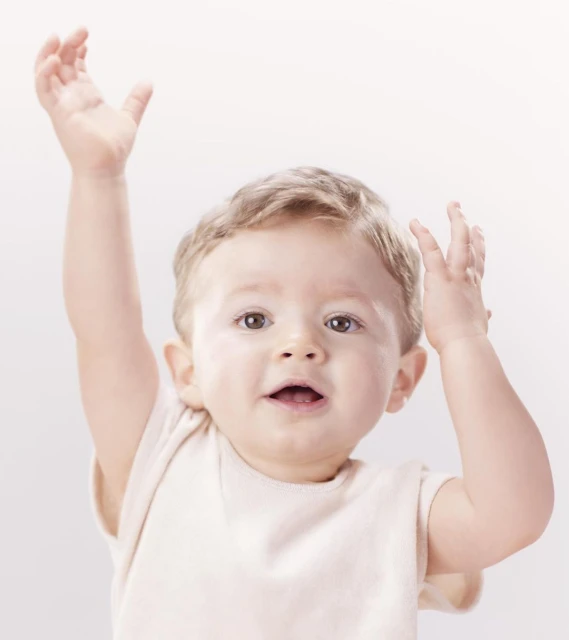 A baby with light brown hair smiles at the camera with arms raised against a neutral background. The baby wears a cream-colored shirt.