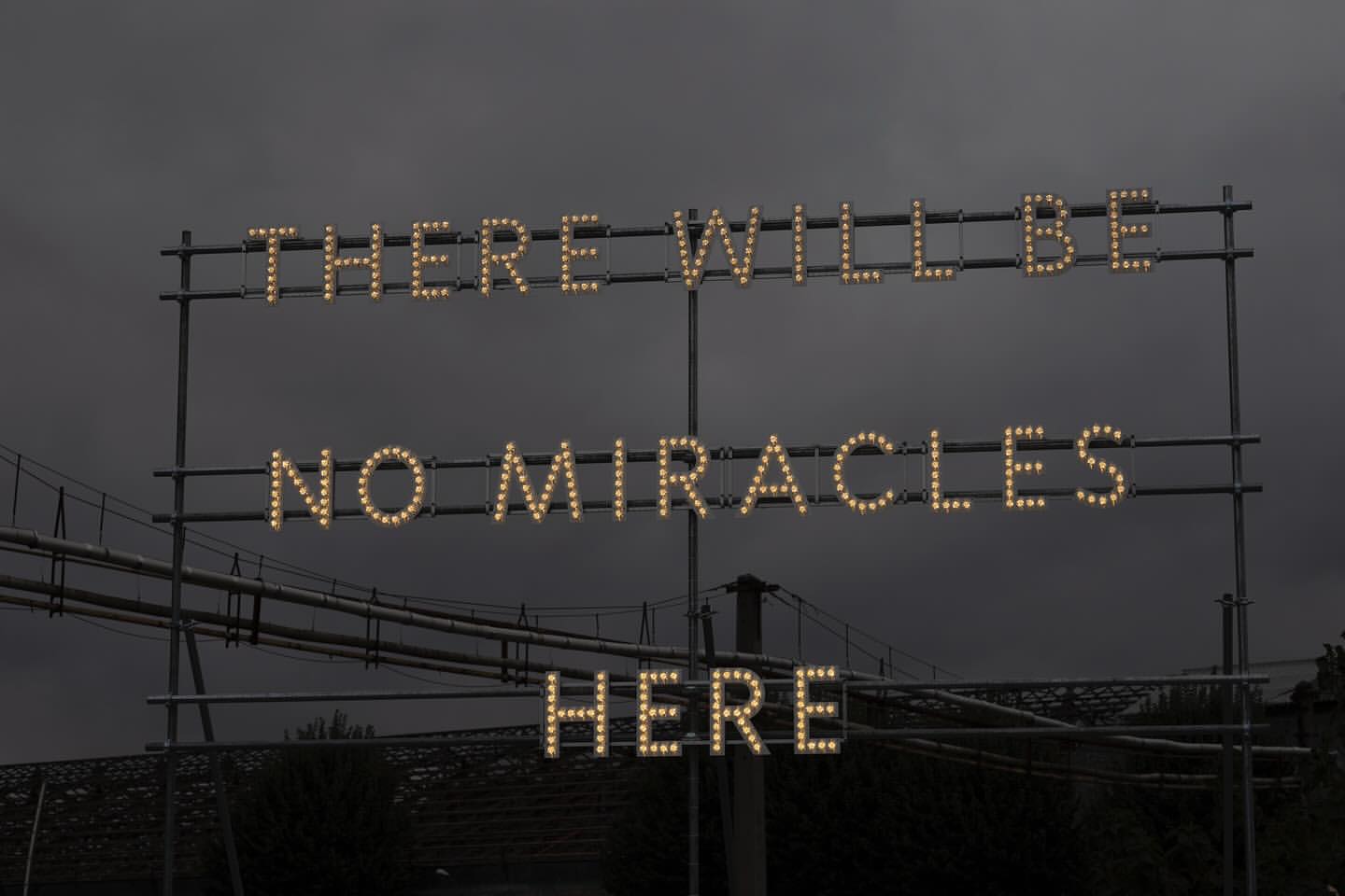 Illuminated sign against a dark sky reads, 'There will be no miracles here,' with a bridge and industrial elements in the background.