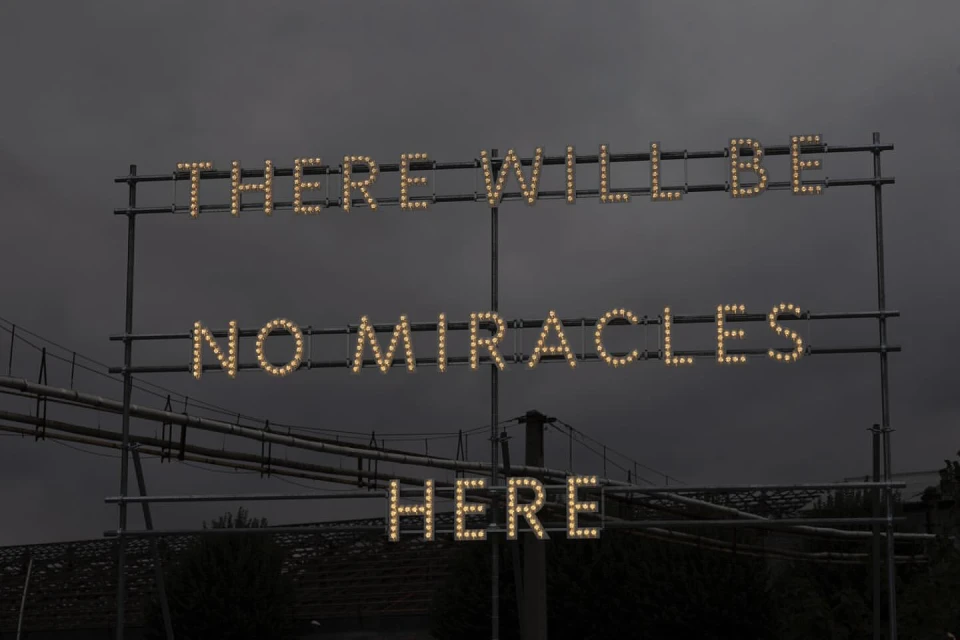 Illuminated sign against a dark sky reads, 'There will be no miracles here,' with a bridge and industrial elements in the background.