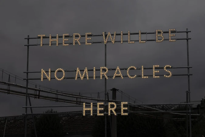 Illuminated sign against a dark sky reads, 'There will be no miracles here,' with a bridge and industrial elements in the background.
