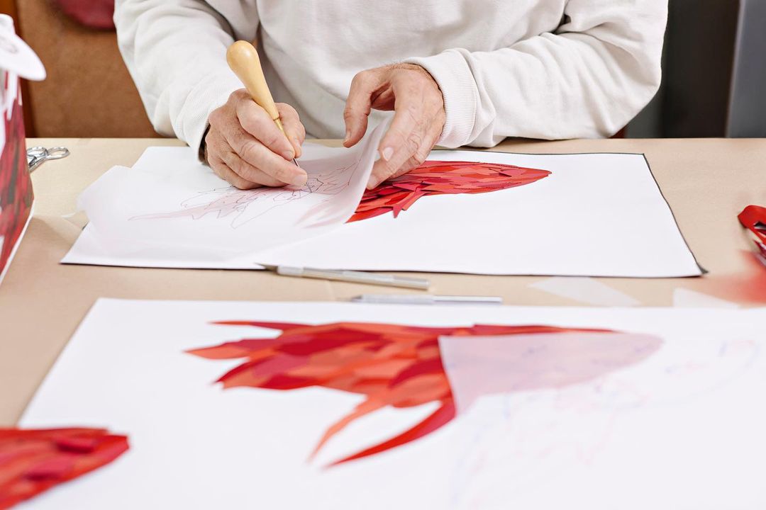 Close-up of an adult's hands using a craft tool to cut out a red mosaic pattern on a white sheet of paper at a table.