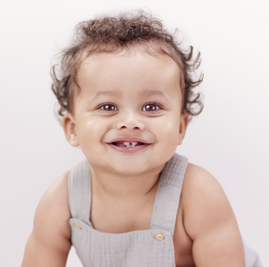 Close-up portrait of a happy baby with curly hair wearing a grey romper, smiling and looking directly at the camera against a white backdrop.