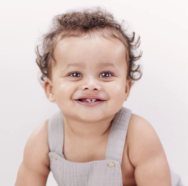 Close-up portrait of a happy baby with curly hair wearing a grey romper, smiling and looking directly at the camera against a white backdrop.