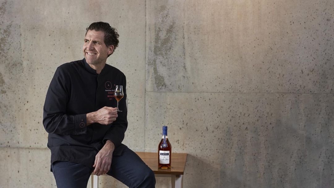 Smiling chef in black jacket seated with a cognac glass next to a Martell bottle, against a textured grey wall indoors.