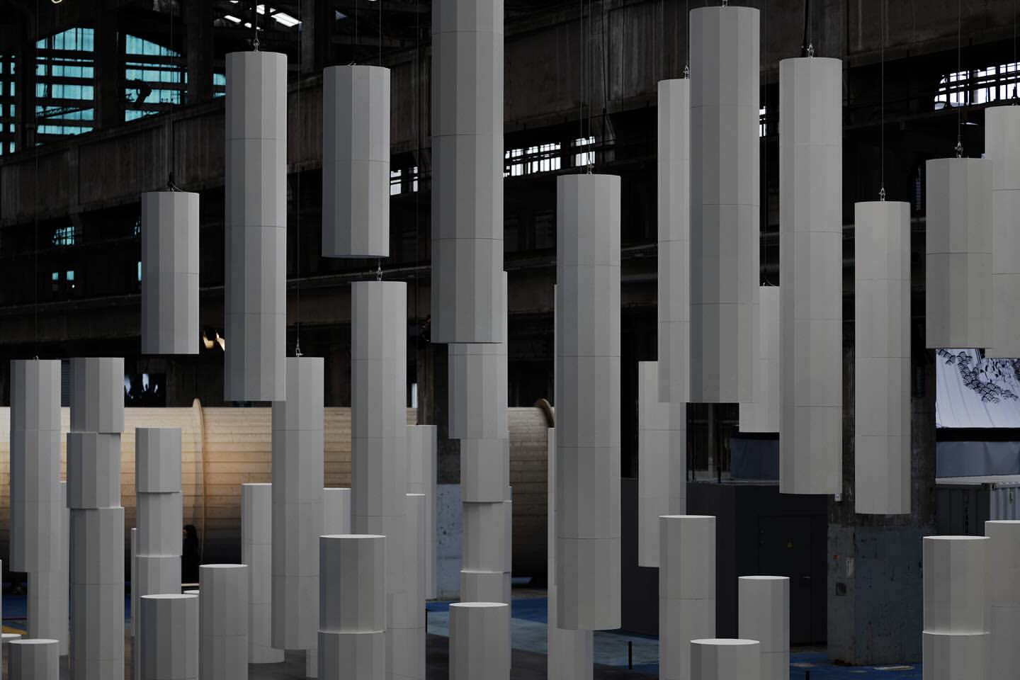 Abstract architectural installation with grey geometric columns hanging from a high ceiling in an industrial building with window light.