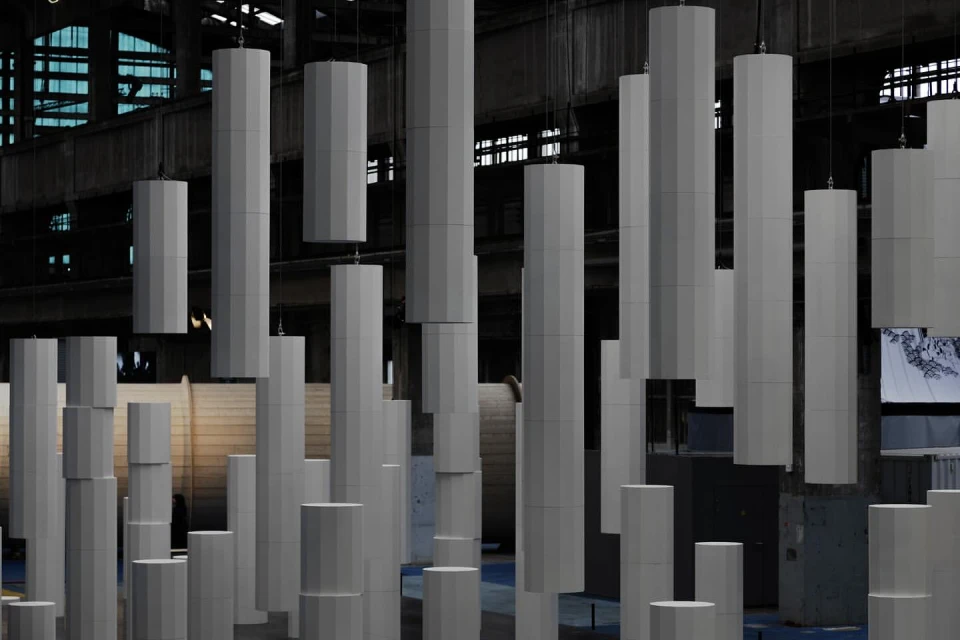 Abstract architectural installation with grey geometric columns hanging from a high ceiling in an industrial building with window light.