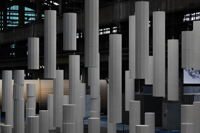 Abstract architectural installation with grey geometric columns hanging from a high ceiling in an industrial building with window light.