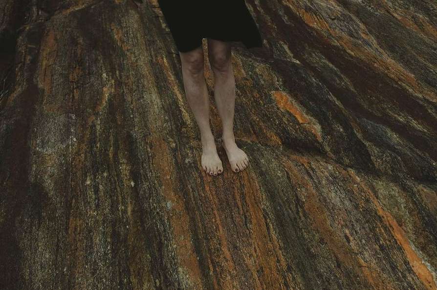 Barefoot woman in a black dress standing on a textured, patterned rock surface with linear formations in shades of brown and orange.