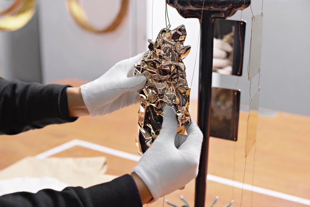 Close-up of gloved hands holding a shiny gold abstract sculpture attached to a metal display stand indoors with a neutral background.