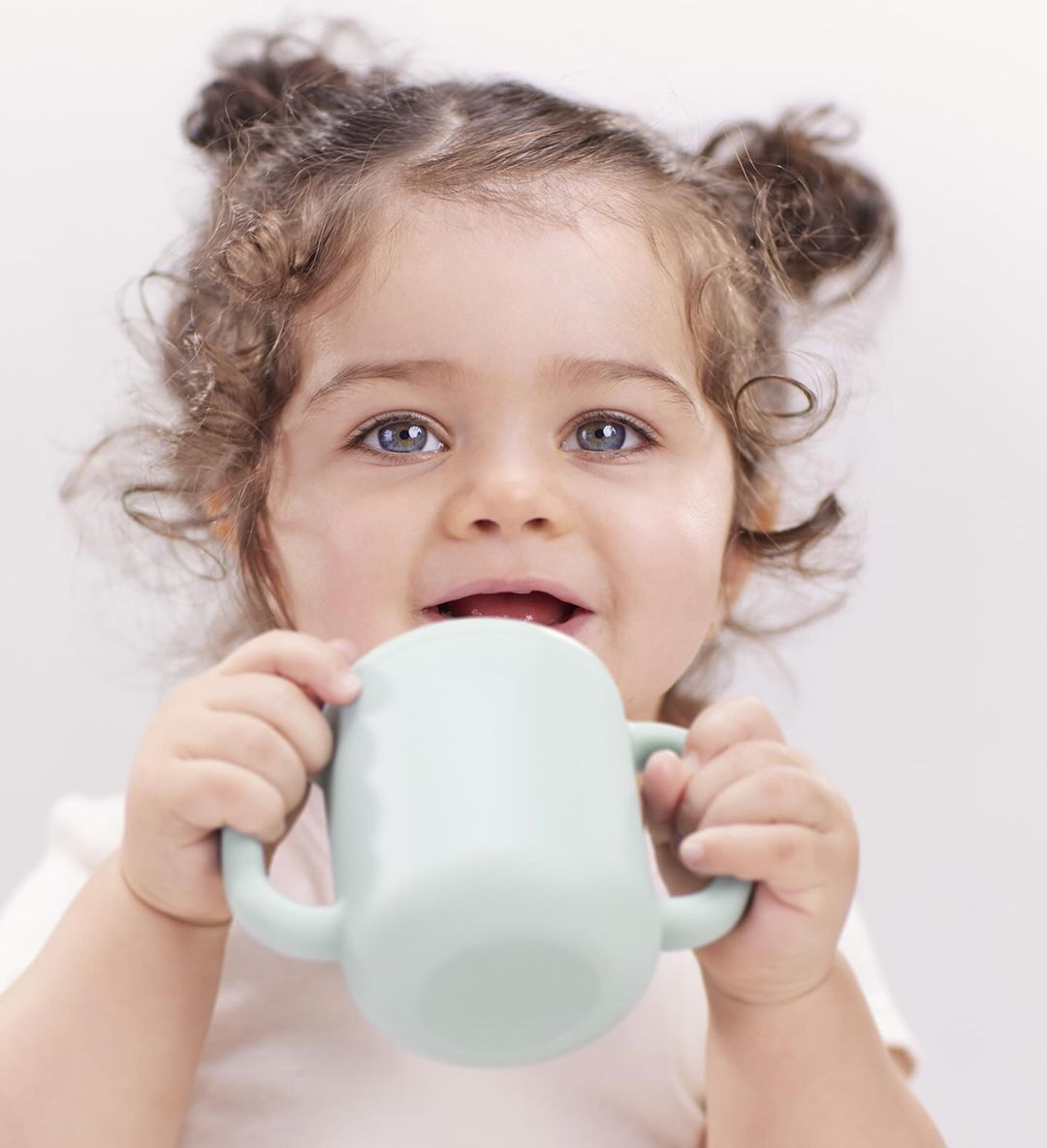 Close-up shot of a child with curly hair drinking from a light green mug with two handles in a brightly lit, white setting.