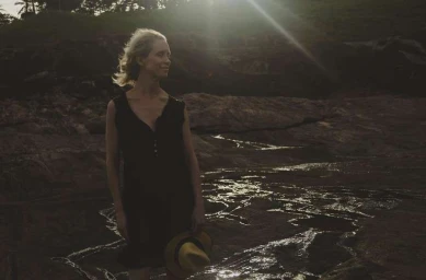 Blonde woman wearing a black dress holding a yellow straw hat standing near rocky terrain at sunset
