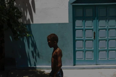 Young boy walking in profile past a blue paneled door and painted wall in a sunlit exterior setting by Jair Lanes.