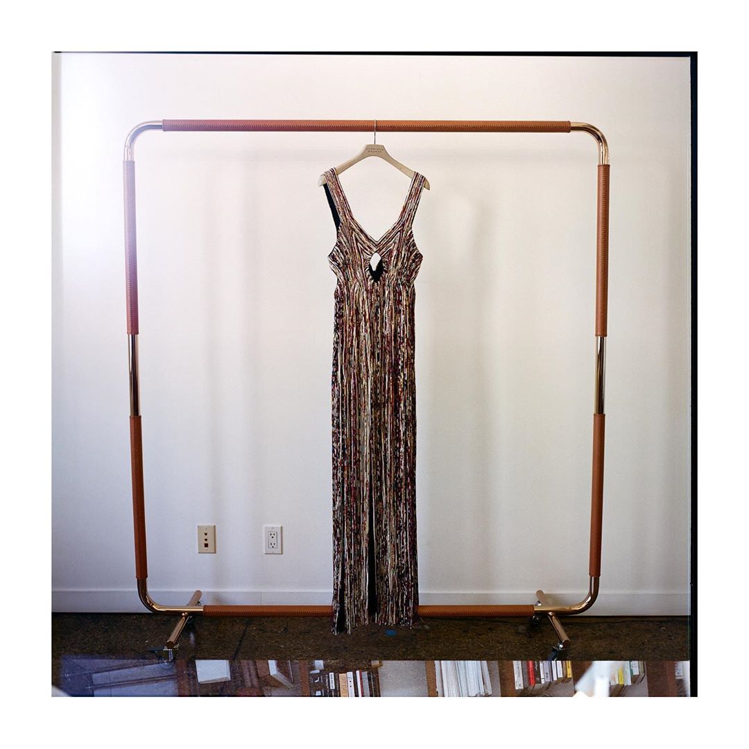 Elegant floor-length dress with a unique pattern hangs on a wood and metal rack against a neutral wall, showcasing fashion in a simple setting.