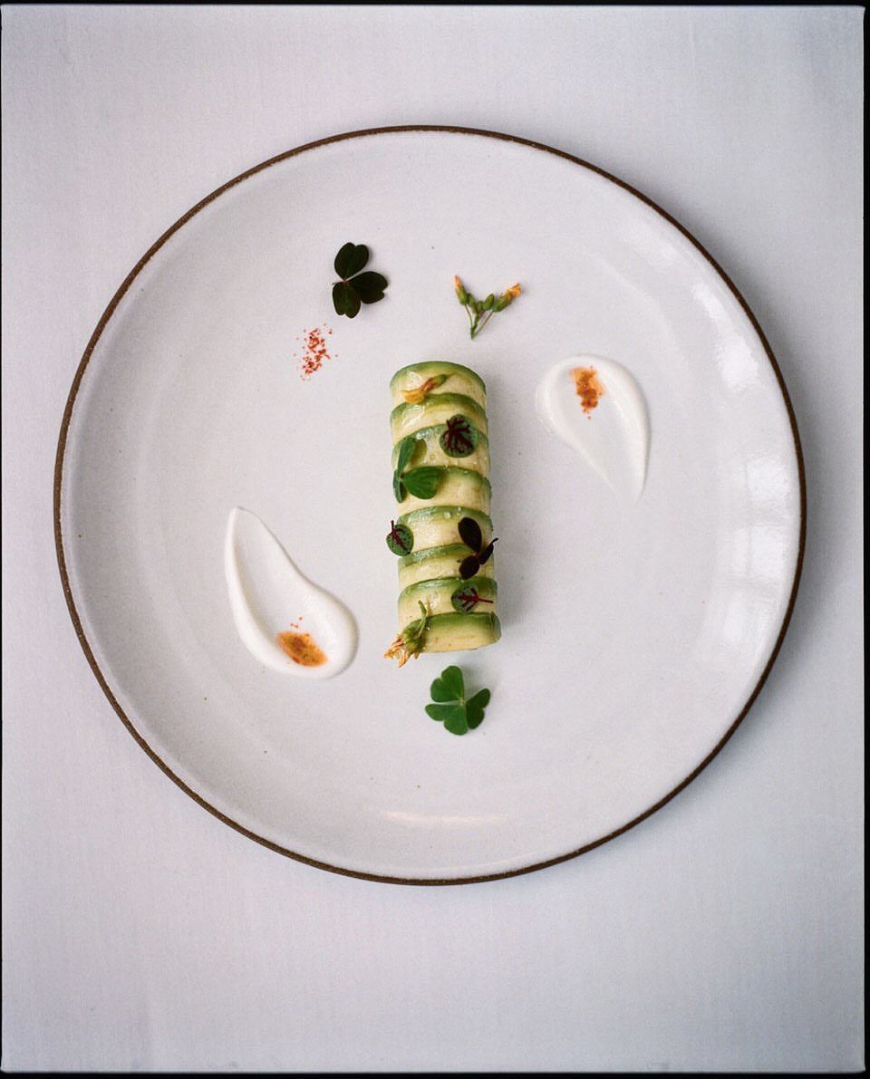 Gourmet food presentation: vertically stacked green zucchini slices with white sauce and green herbs on a round white plate.