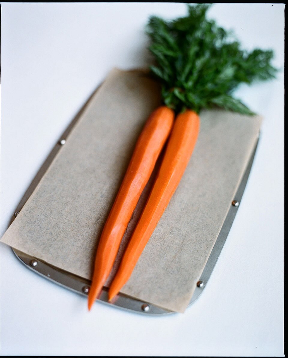 Two fresh carrots with green leafy tops lie side by side on parchment paper on a metal baking sheet, on a white surface.