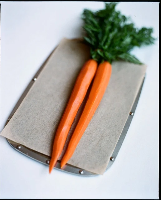 Two fresh carrots with green leafy tops lie side by side on parchment paper on a metal baking sheet, on a white surface.