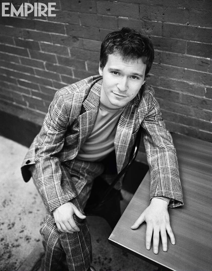 Black and white portrait of a man in a plaid suit sitting next to a table, posing for a photo in front of a brick wall.