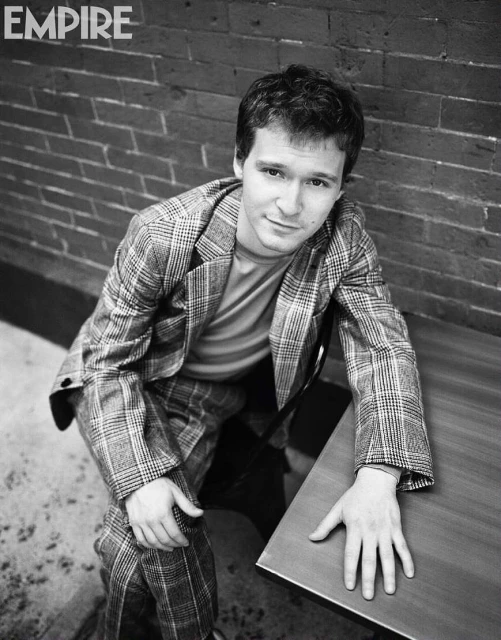 Black and white portrait of a man in a plaid suit sitting next to a table, posing for a photo in front of a brick wall.