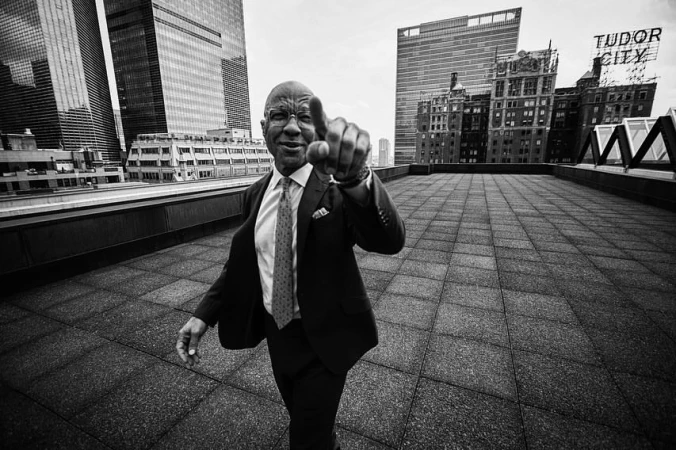 Black and white portrait of a man in a suit pointing towards the viewer on a rooftop with city buildings in the background.
