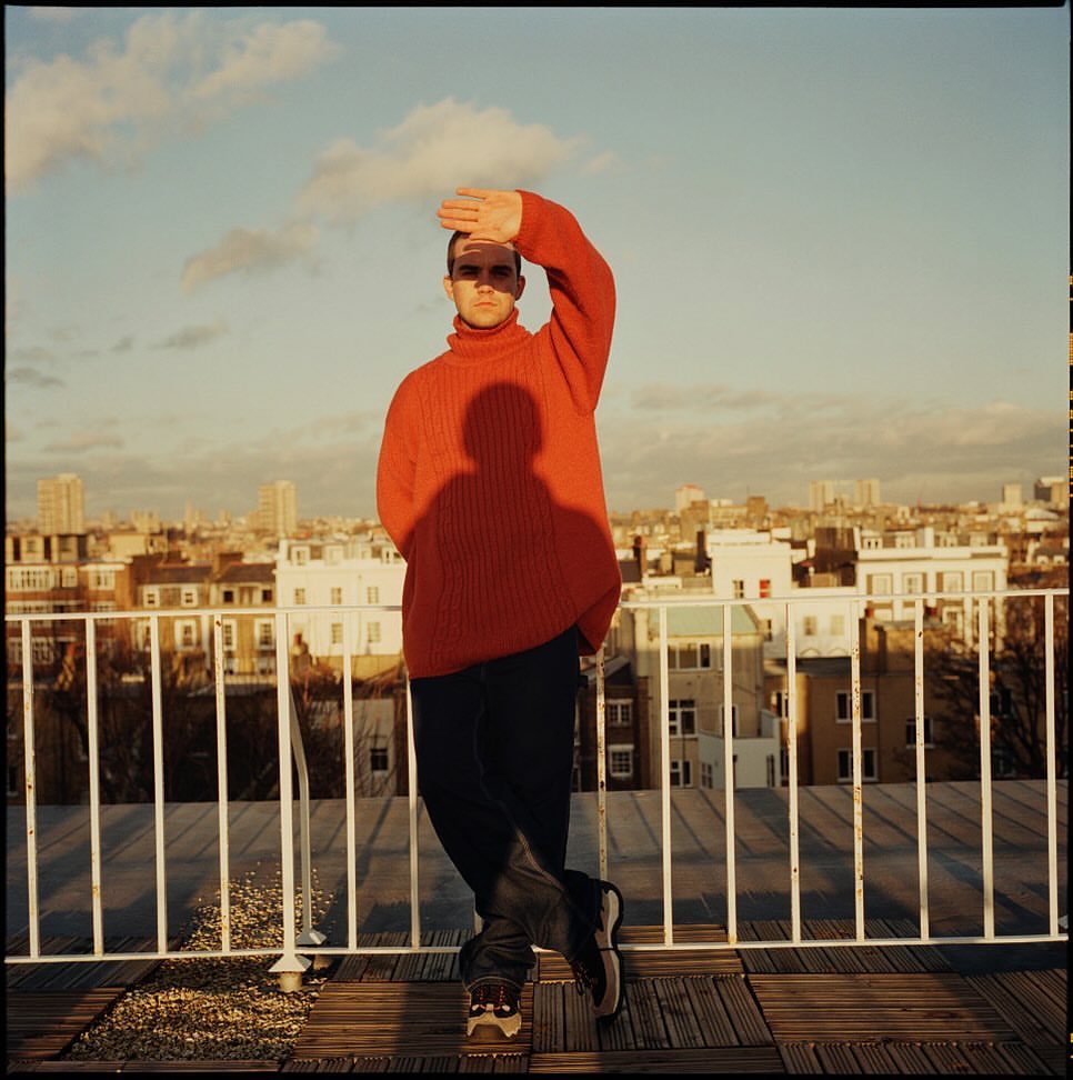 Man in orange sweater shields eyes on rooftop with city view backdrop; sunlight, shadows, casual clothes, outdoor setting, relaxed pose.